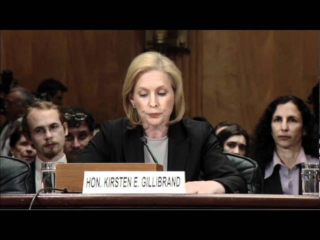 Blonde woman in suit at a podium with microphones, nameplate reading “HON. KIRSTEN E. GILLIBRAND.”