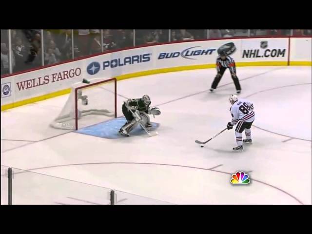 Hockey goalie crouches in front of the net as a skater and referee stand nearby.