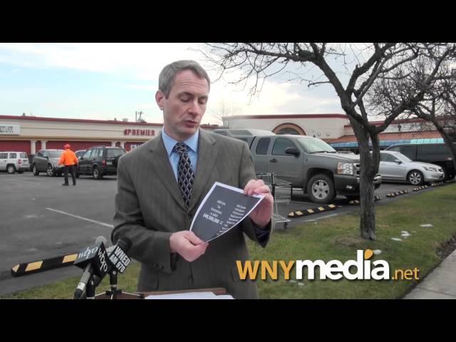 Reporter in suit holding a folder and speaking outdoors near parked vehicles