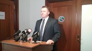 Speaker at a podium behind several microphones, indoors near a door and wall logo