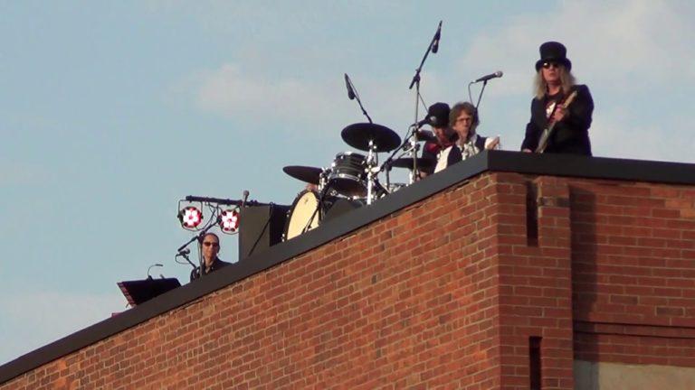 Musicians in dark outfits play on a rooftop stage with drums and microphones