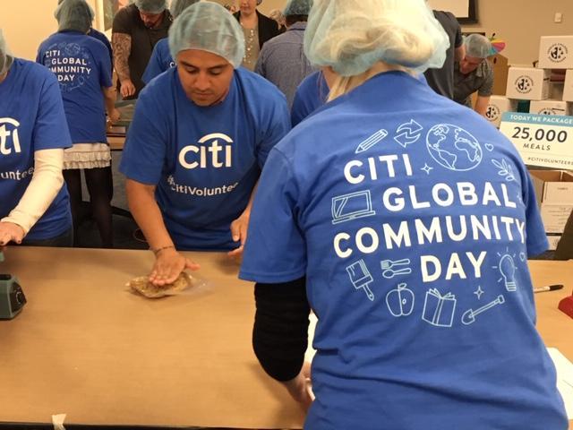Volunteers in blue Citi Global Community Day shirts assemble meal packages at a table.
