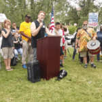 Person speaking at a podium outdoors as a crowd holds signs and banners.