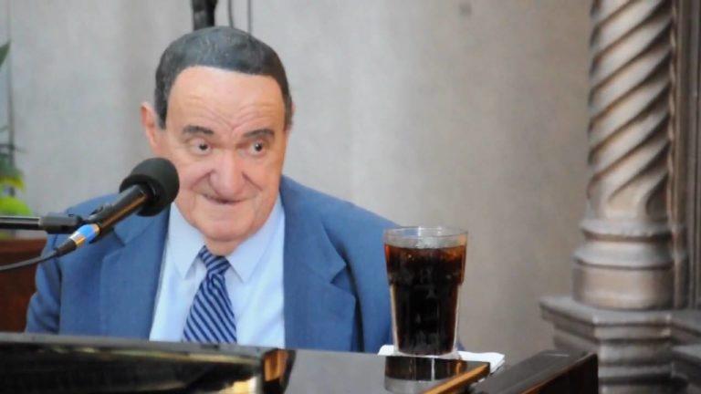 Man in a blue suit speaks into a microphone; a glass of dark drink sits on the table.