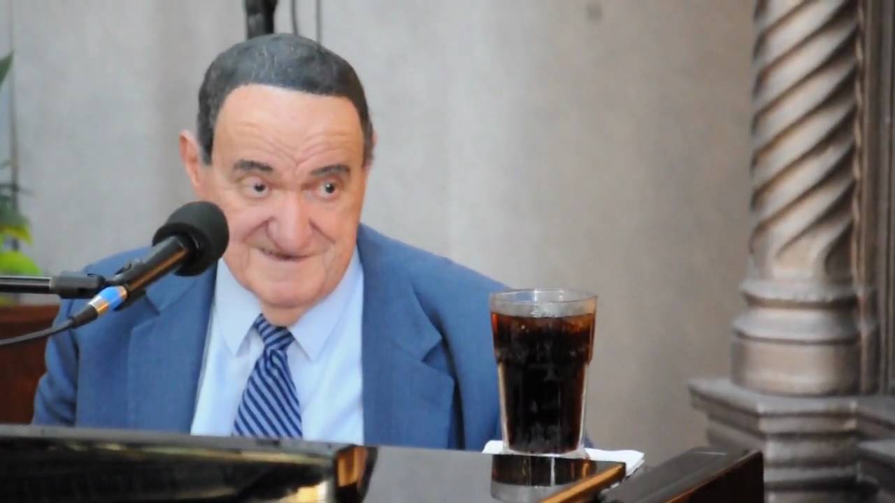 Man in a blue suit speaks into a microphone; a glass of dark drink sits on the table.