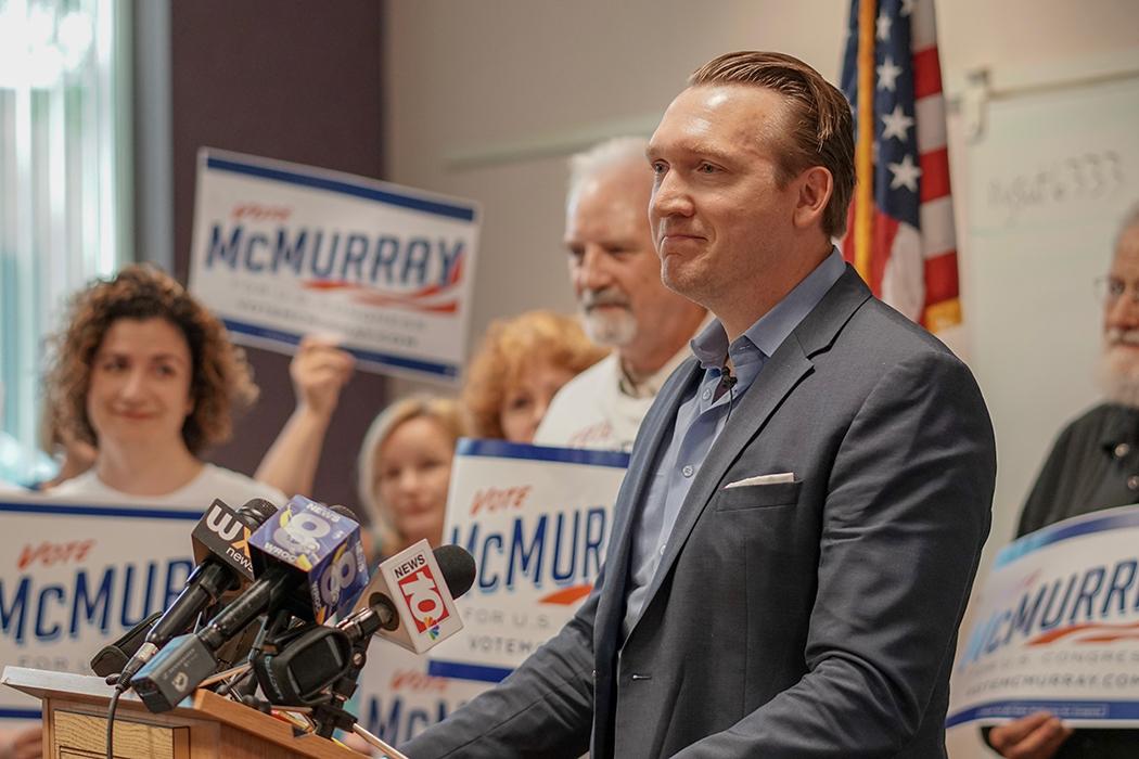 Man in suit speaks at a podium with multiple microphones during a campaign event.