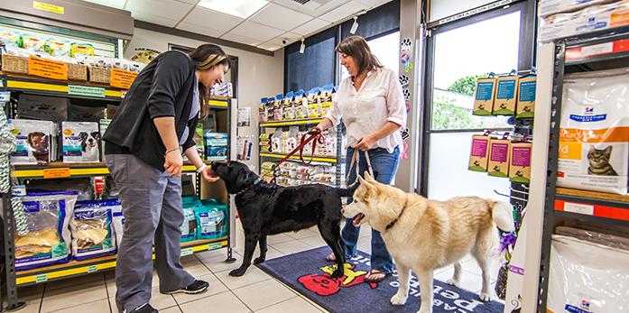 Two people stand with two dogs in a pet store aisle near shelves of pet supplies.
