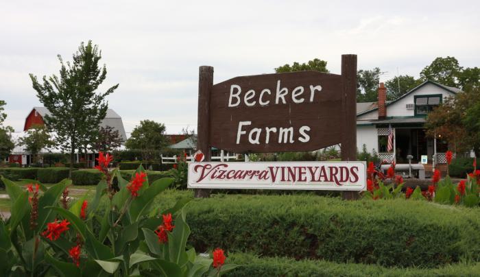 Wooden sign reading “Becker Farms” with “Vizcarra VINEYARDS” banner in front of buildings and greenery.