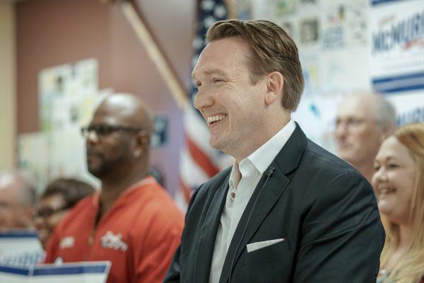 Man in a dark suit smiles and speaks while people sit and stand behind him