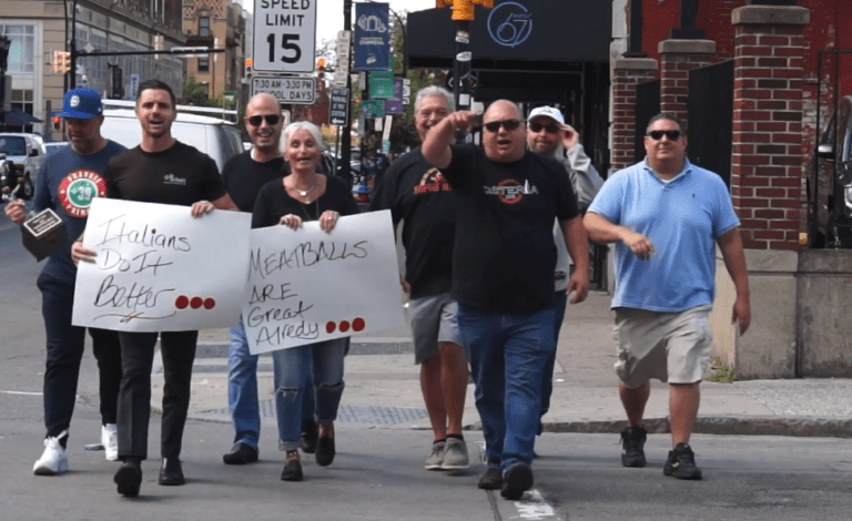 People walk on a street holding handwritten protest signs with black-and-red text.