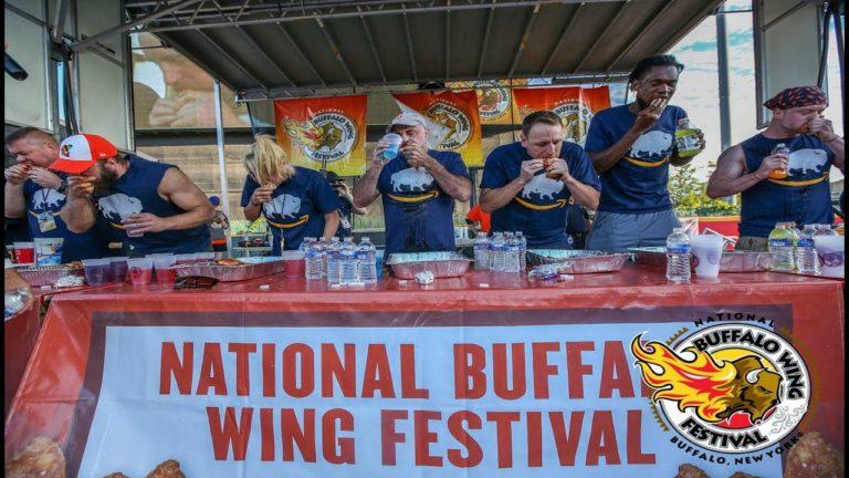 Attendees in team shirts eat wings at a festival buffet table under a tent.