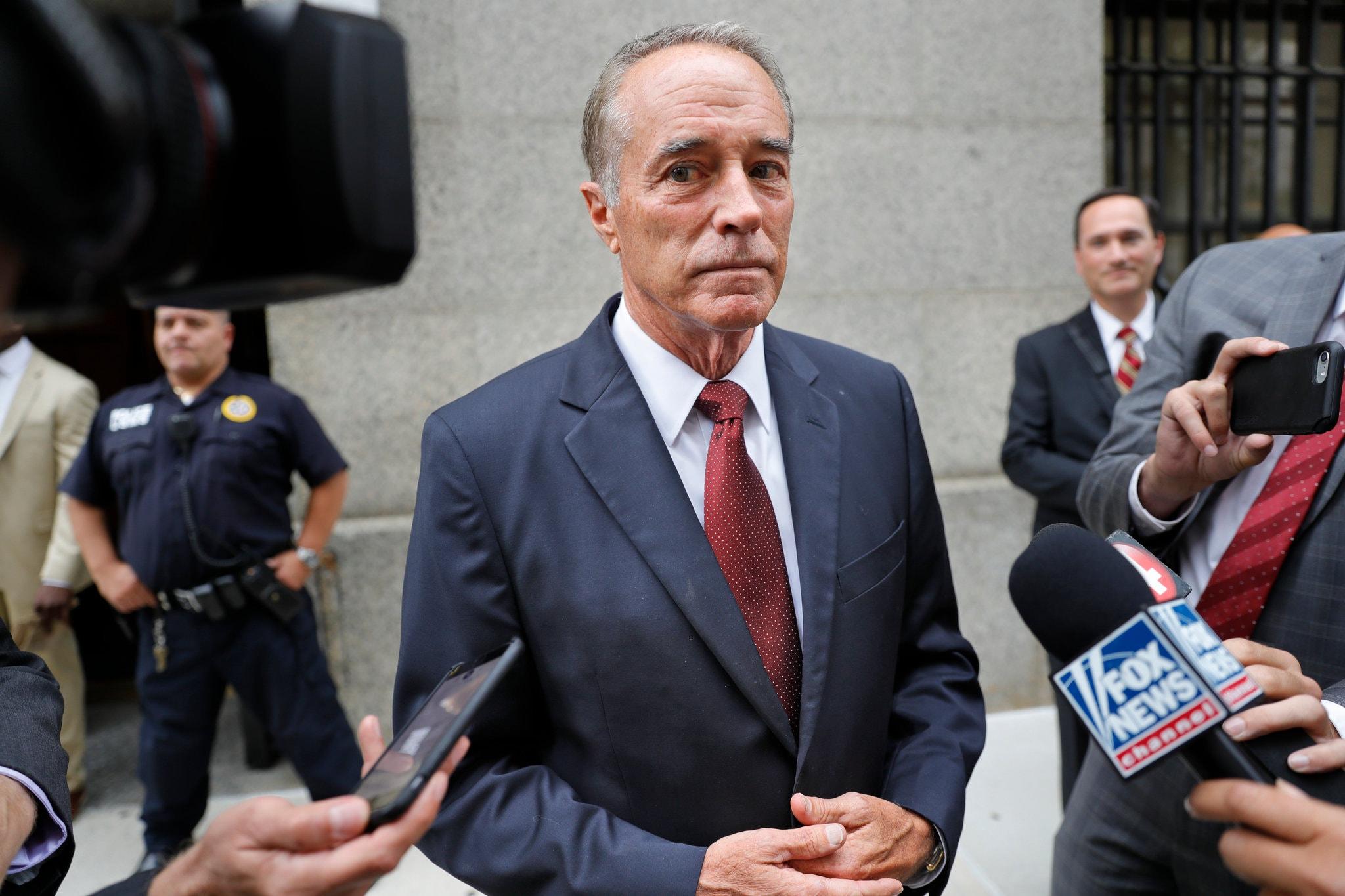 Man in a dark suit and red tie stands outside as reporters hold microphones and cameras.