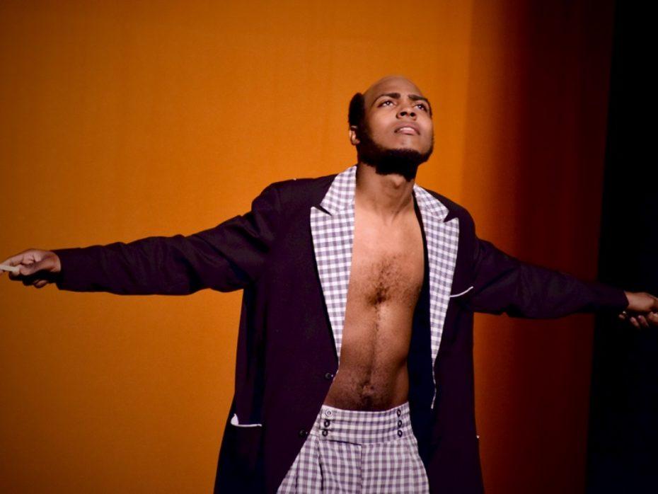 Man in an open jacket and plaid pants stands on stage with arms extended.