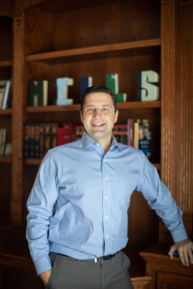Smiling man standing in front of a wooden bookshelf with books behind him.