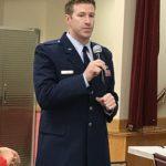 Uniformed person speaking into a handheld microphone in a room with red curtains