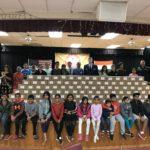 Group of adults and children posing in front of stacked cardboard boxes in a hall.