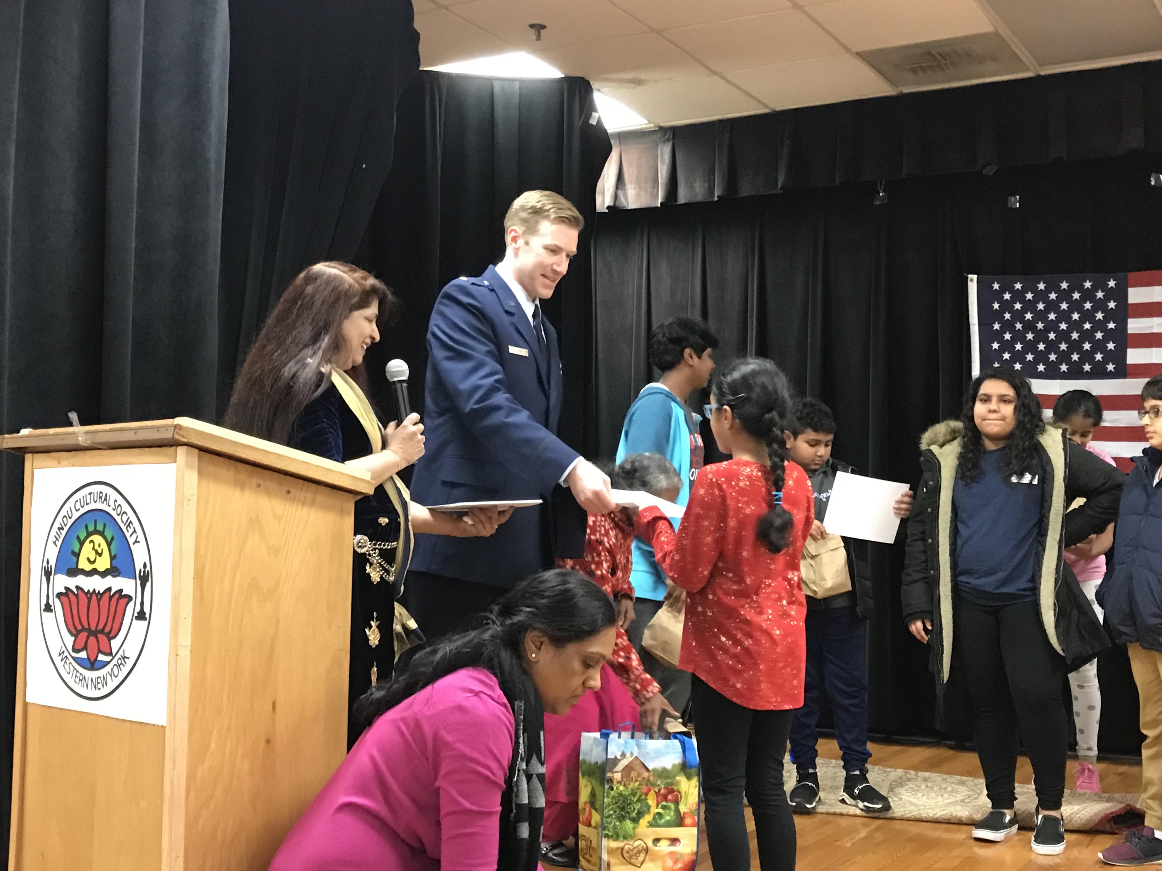 Speaker in blazer hands items to children near a podium with a Western New York logo.