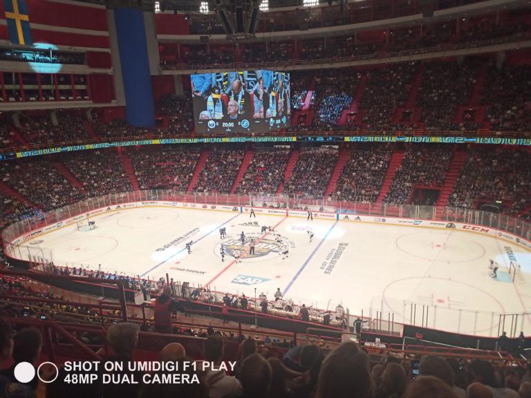 Indoor hockey arena viewed from stands with players on the ice and a scoreboard screen.