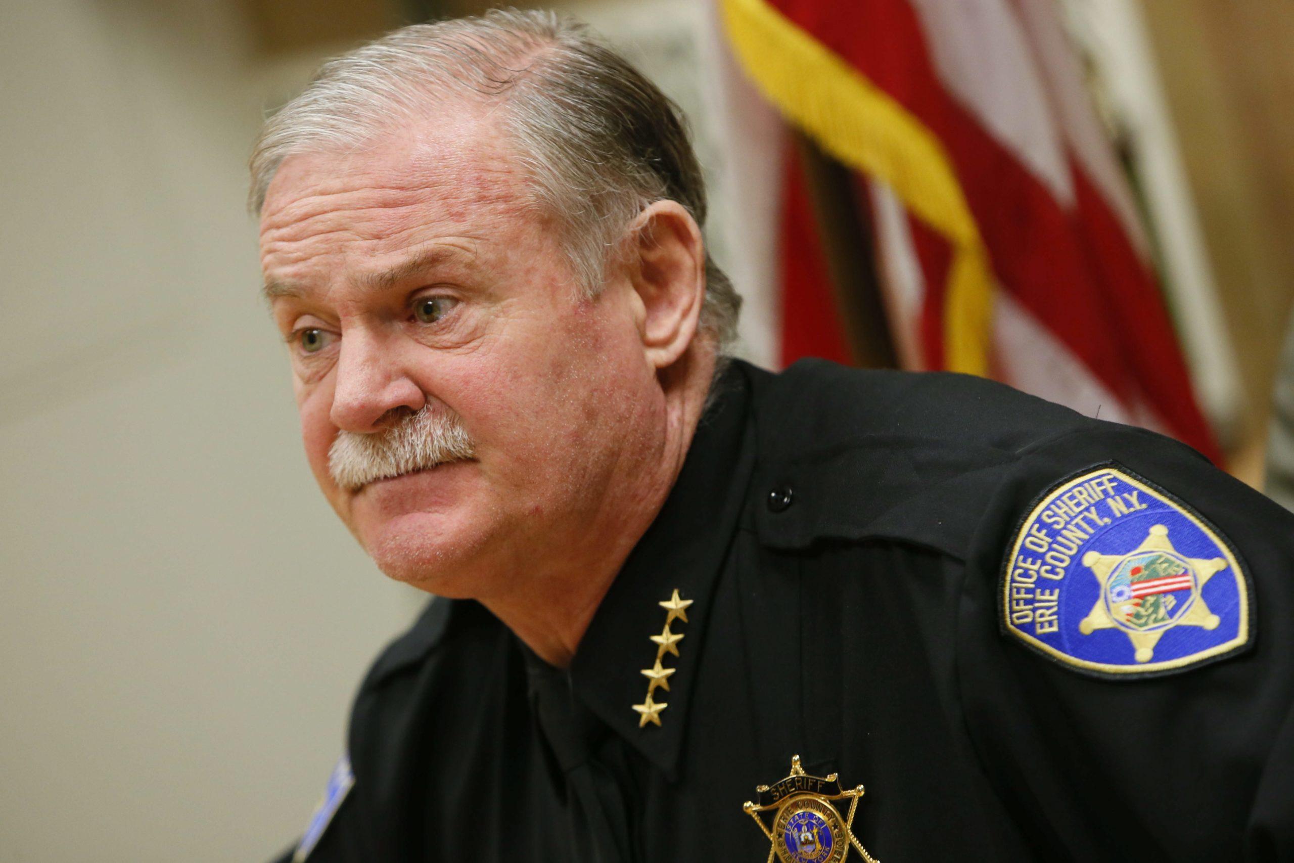 Man in dark sheriff uniform with a badge patch and star insignia against blurred flags