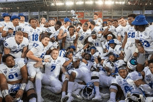 Football players and coaches huddle and celebrate on a stadium field under bright lights.
