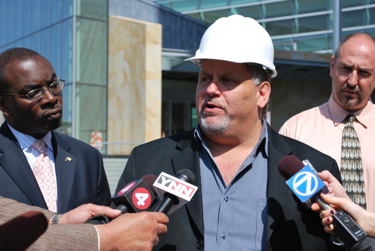 Man in white hard hat speaks at microphones while reporters hold recording devices nearby