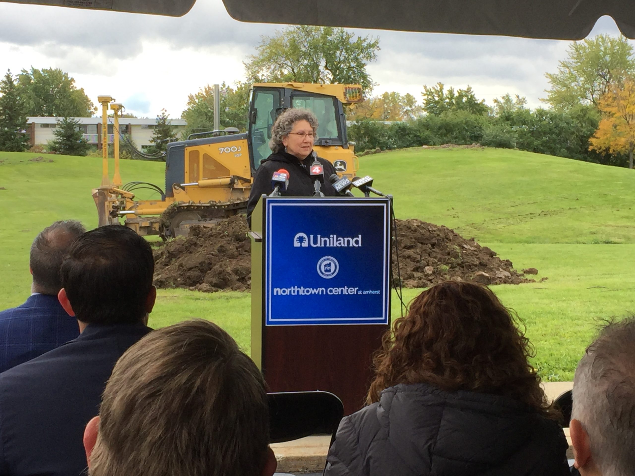 Spokesperson at a blue podium with microphones in front of a bulldozer and lawn.
