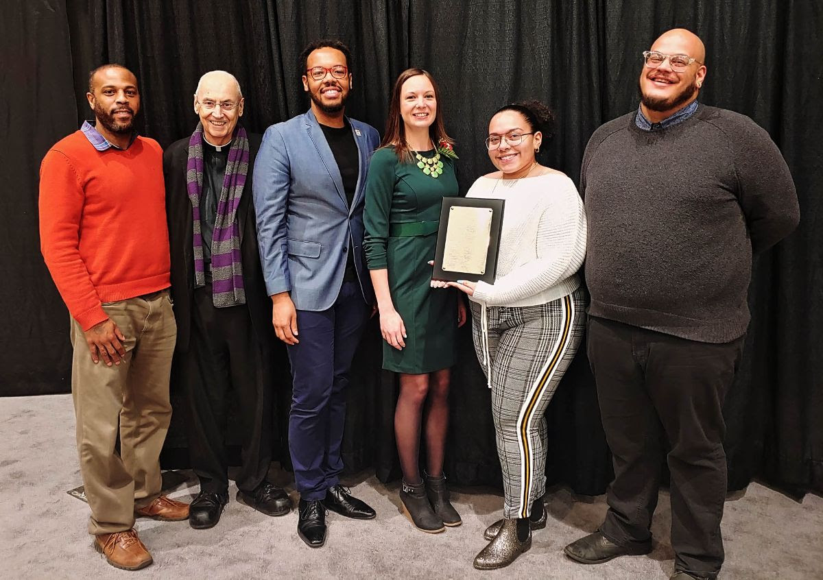 Five people pose in front of a curtain; one holds a framed certificate, others stand smiling.