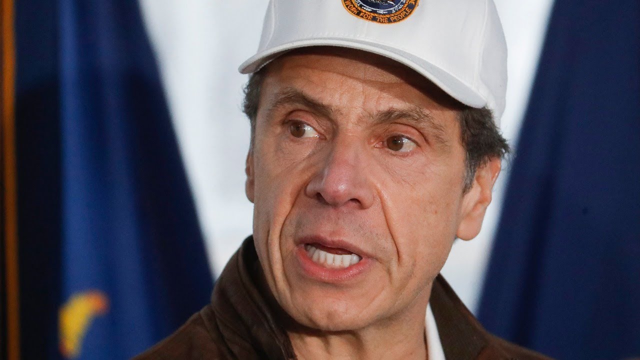 Older man wearing a white cap speaks in front of flags and a microphone background.