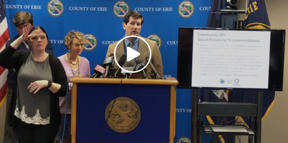 Officials stand at a podium with microphones and a monitor showing social distancing recommendations.