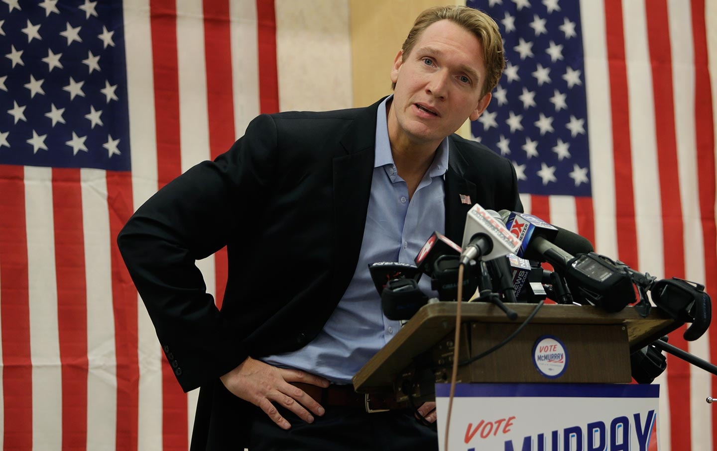 Person in a suit speaks at a podium with multiple microphones in front of U.S. flags.