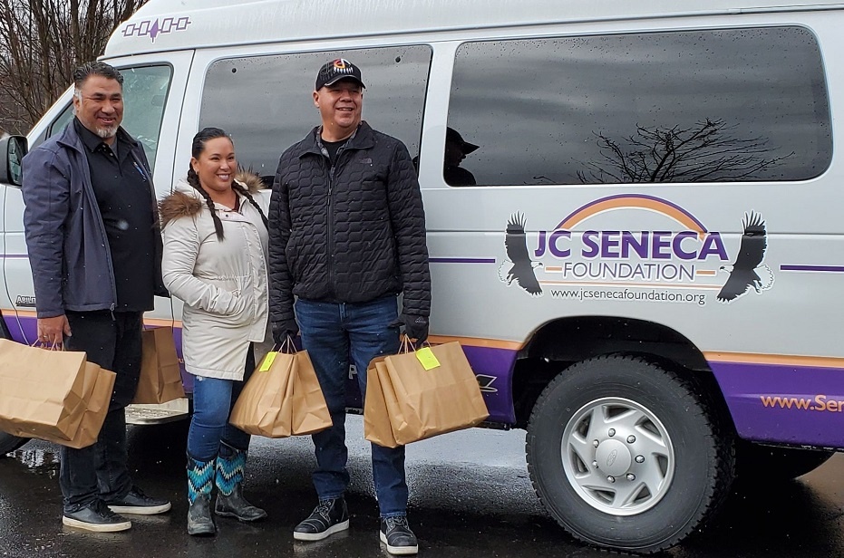 Three people pose with paper gift bags in front of a JC Seneca Foundation van.