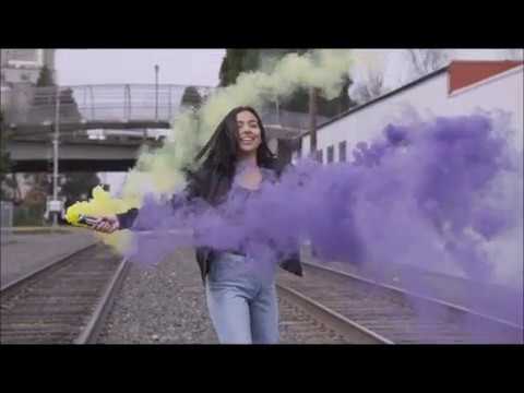 Person holding a yellow electric guitar in front of purple and green smoke near railway tracks