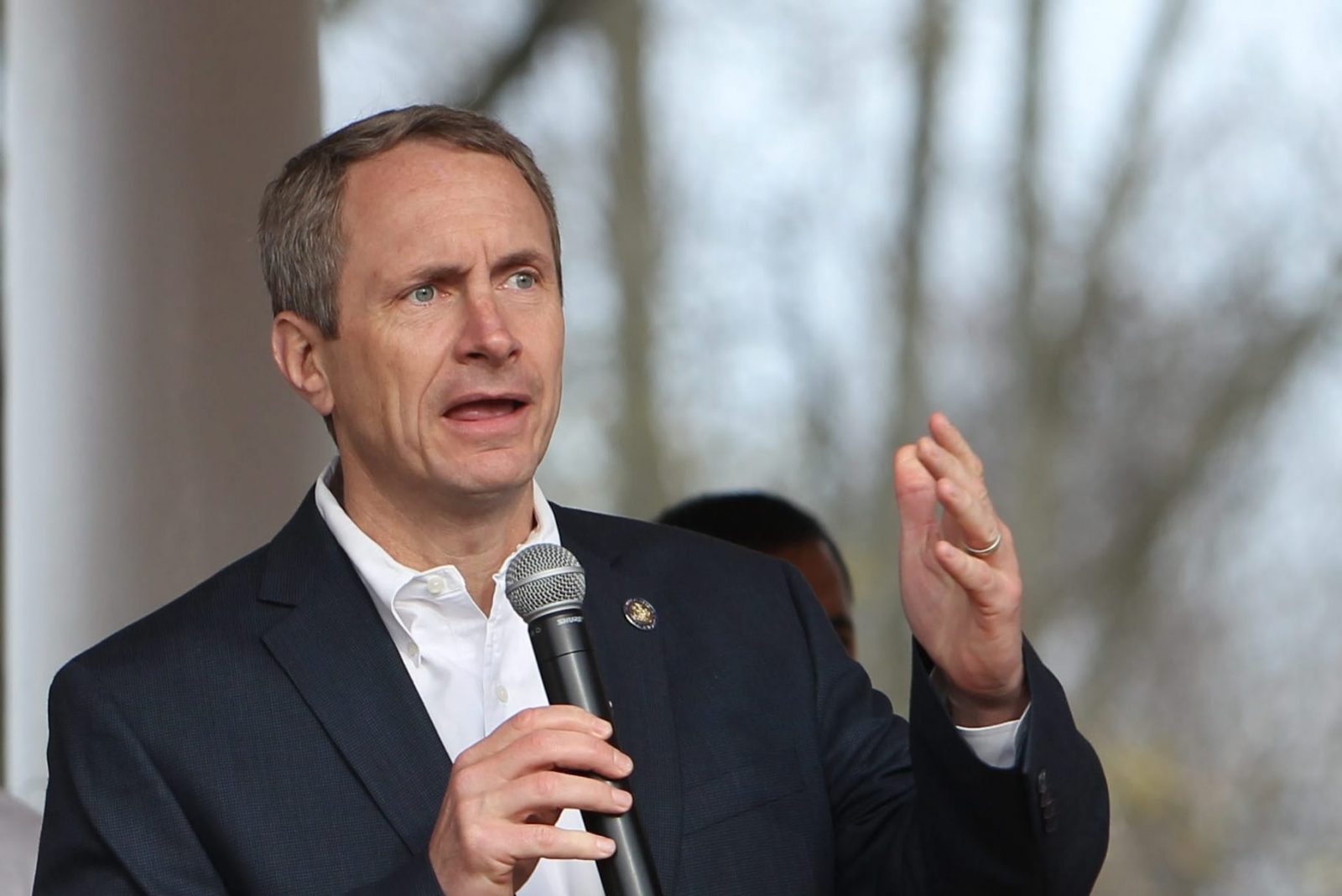 Man in a suit speaking into a handheld microphone, raising his hand outdoors.