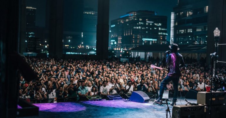 Performer stands on a stage with microphones and equipment while a large crowd watches at night.