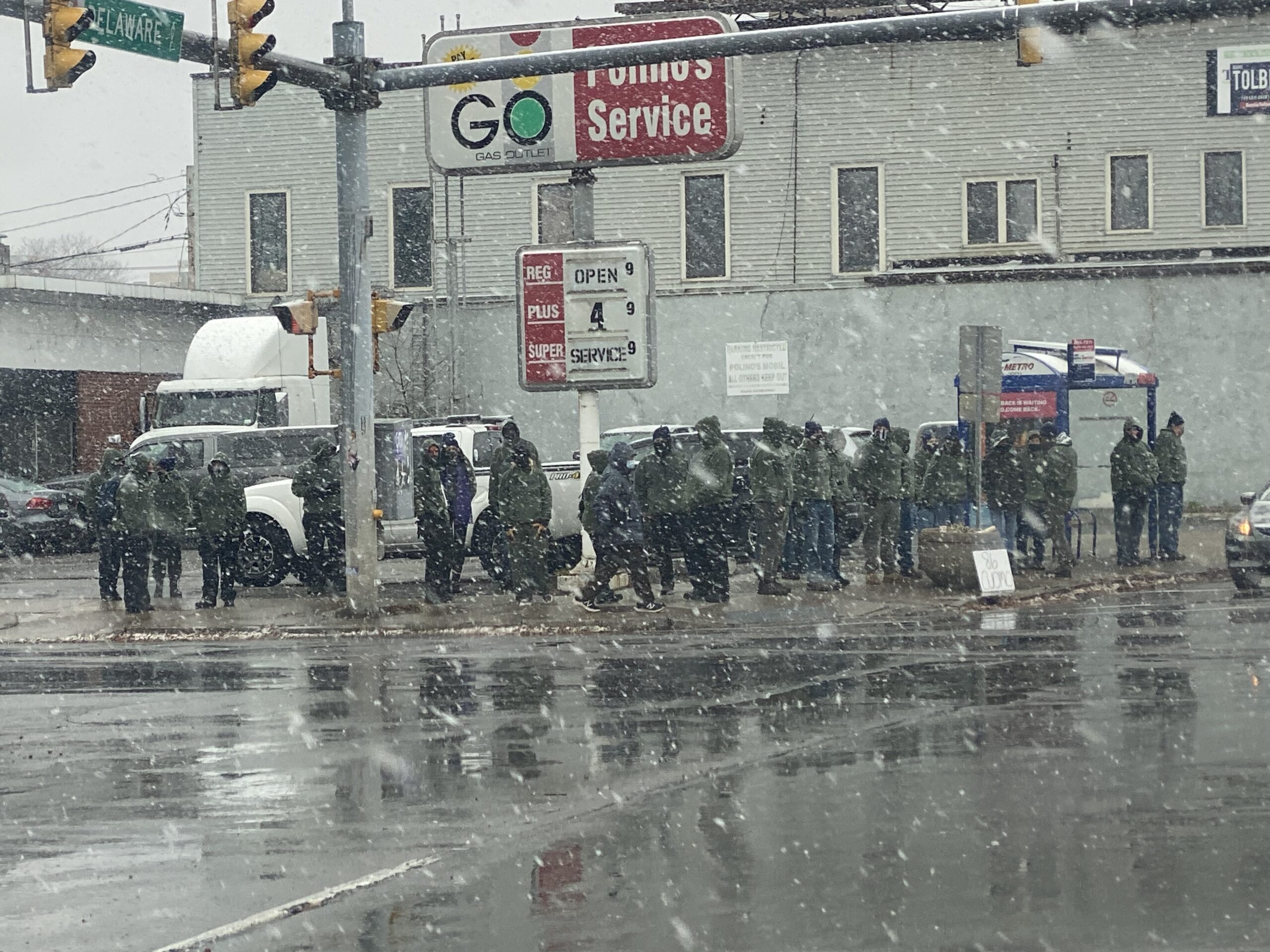 People in uniforms stand in snowy street near a storefront and parked vehicles.