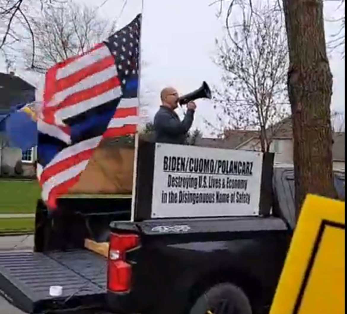 Man speaks at a podium with a megaphone beside a large U.S. flag on a trailer.