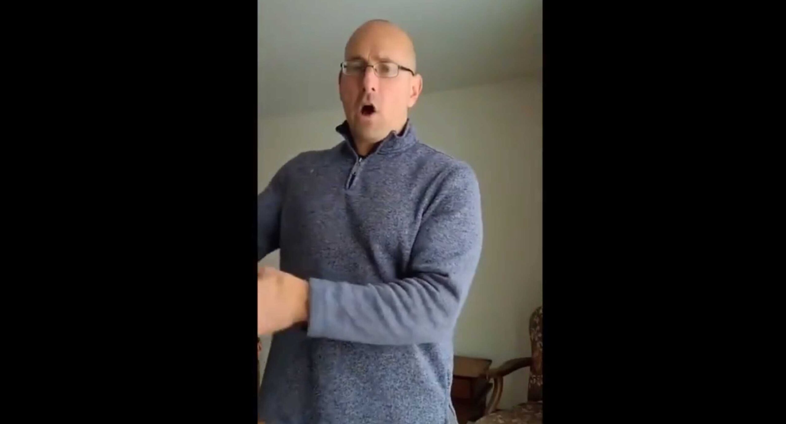 Bald man wearing glasses in a gray sweater gestures while speaking indoors.