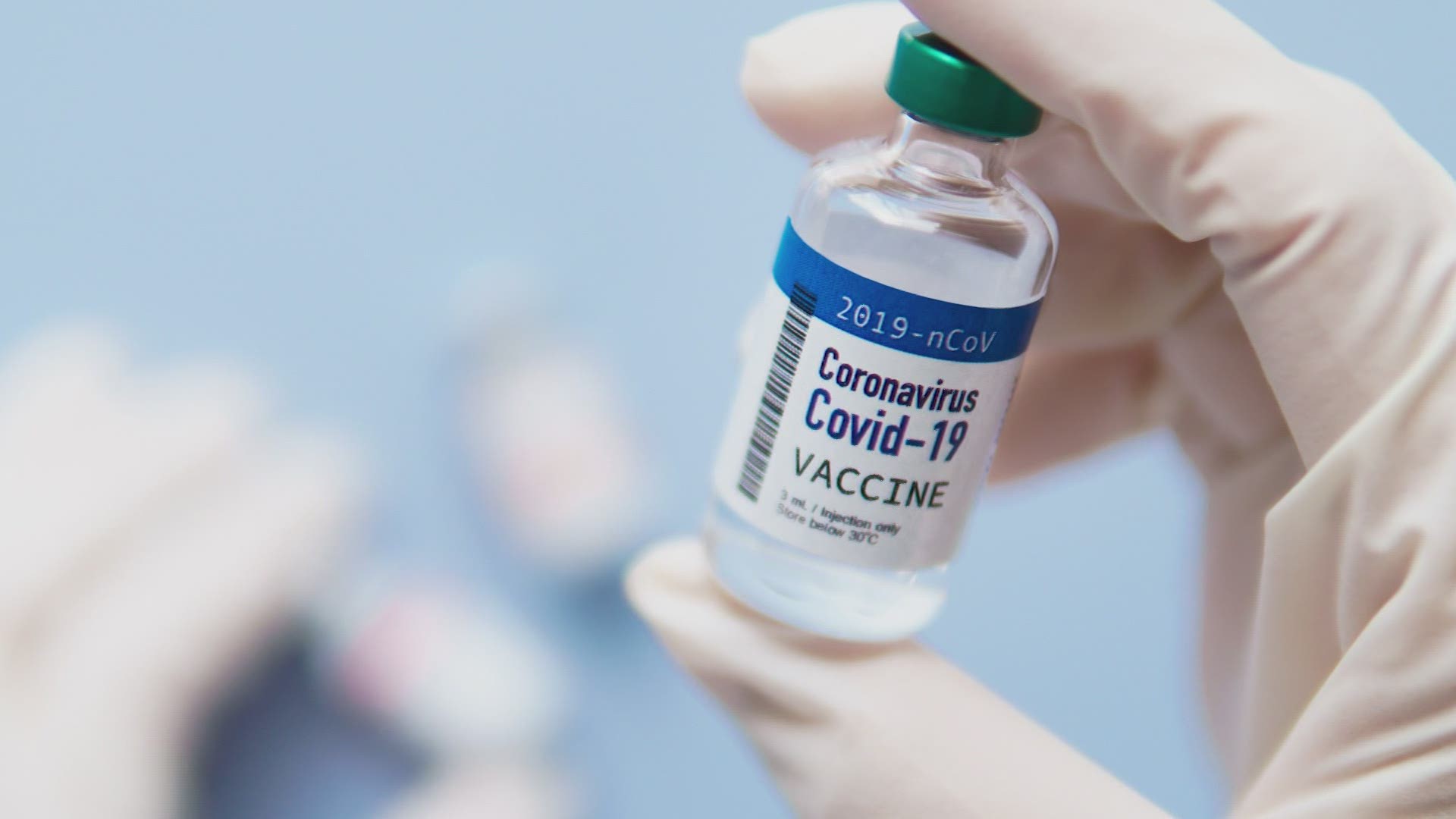 Gloved hand holds a labeled Covid-19 vaccine vial against a blurred background.