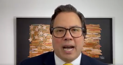 Person wearing glasses speaking at a desk with shelves of books behind them
