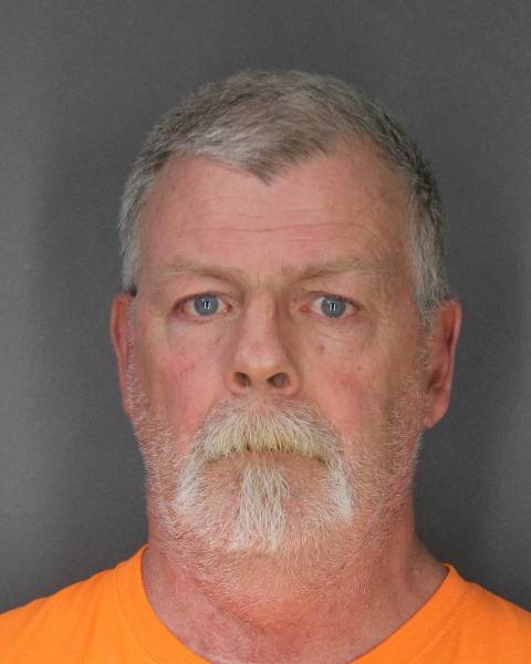 Man with gray hair and goatee wearing an orange prison shirt against a gray background.