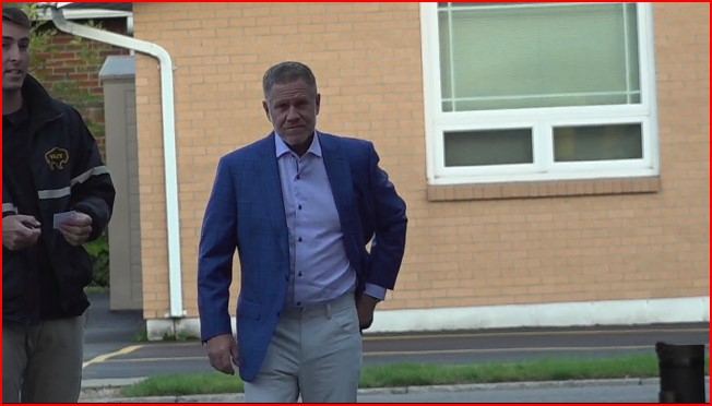 Man in a blue blazer walking outside a brick building with a window behind him