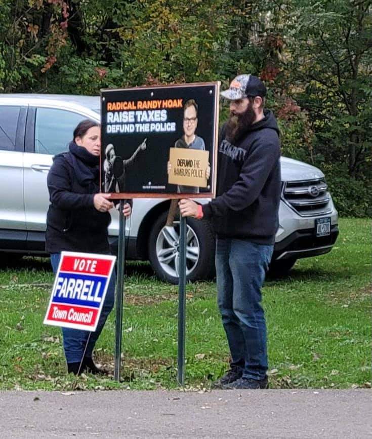 Two people stand by campaign signs on a grassy roadside near parked cars.