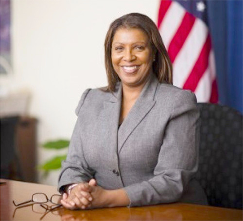 Smiling woman in a gray blazer seated at a desk with U.S. flags behind her