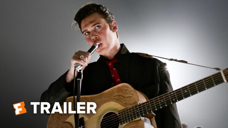Performer sings into a microphone while holding an acoustic guitar against a gray backdrop.