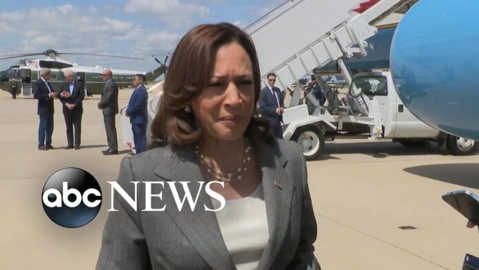 Woman in a gray blazer stands in front of an airport aircraft and microphones.