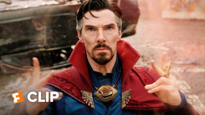 A man in a red cloak and blue shirt looks at the camera with hands raised.