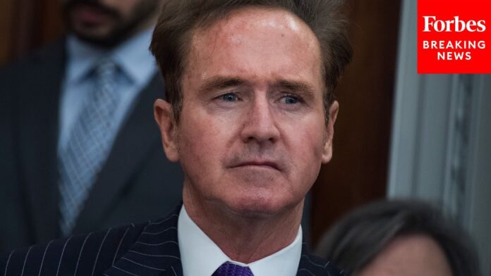 Man in a dark suit and purple tie with a red “Forbes Breaking News” label in the corner.