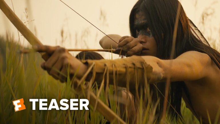 Woman with long hair crouches in tall grass, hands gripping another person’s arm.