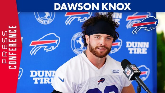 Football player in a white jersey and cap at a podium with a team logo backdrop