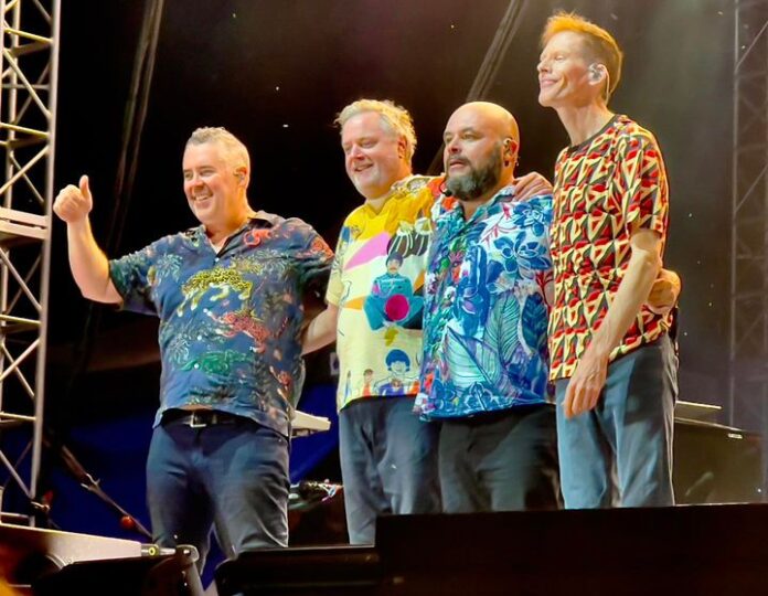 Four men in colorful patterned shirts stand onstage, smiling and posing.